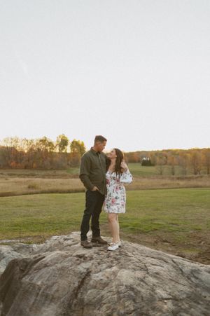 A couple stands together on a large rock formation during golden hour in a rural landscape.