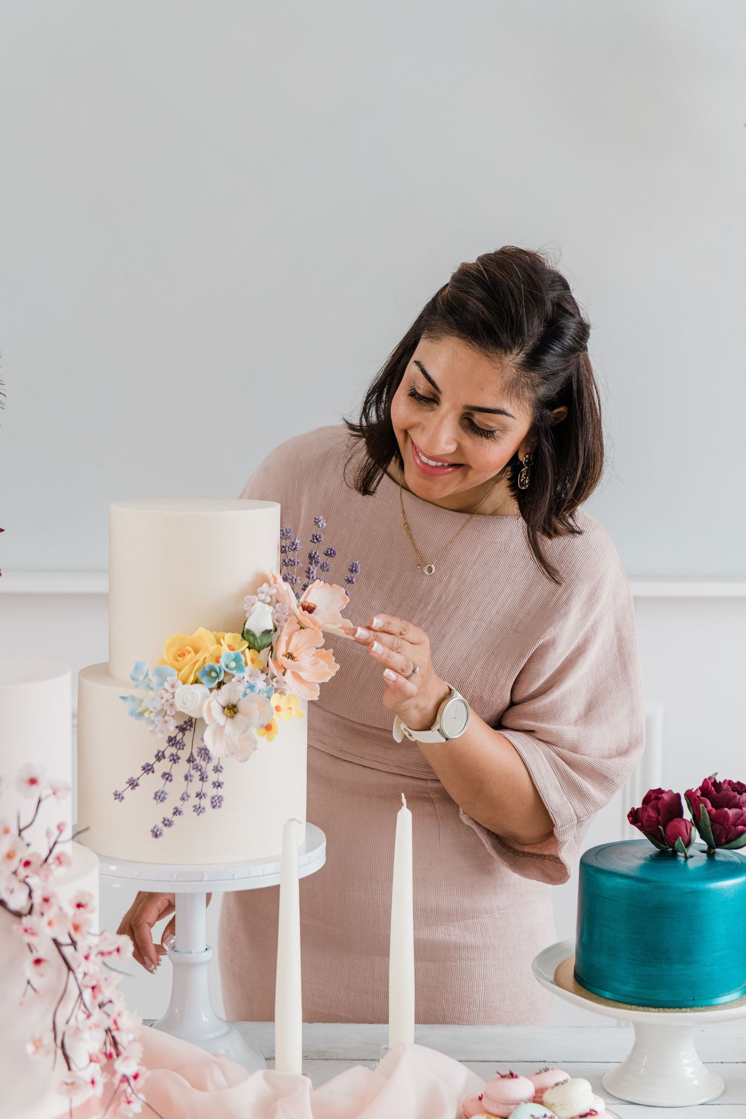 Baker decorating white and teal wedding cakes with delicate floral details on cake stands against neutral background.