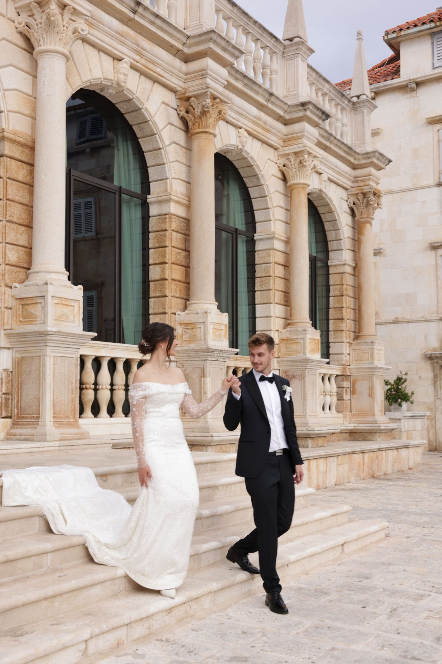 A couple in wedding attire playfully runs through an elegant stone archway corridor with classical architecture.
