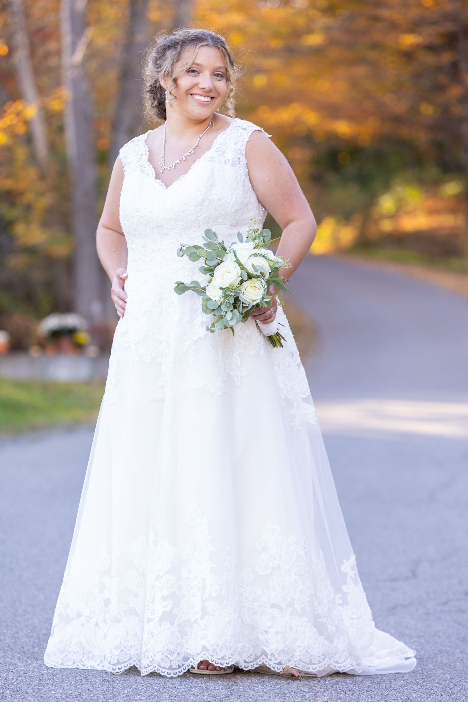 A bride in a beautiful lace-trimmed white wedding gown holds a white and green bouquet against autumn foliage.