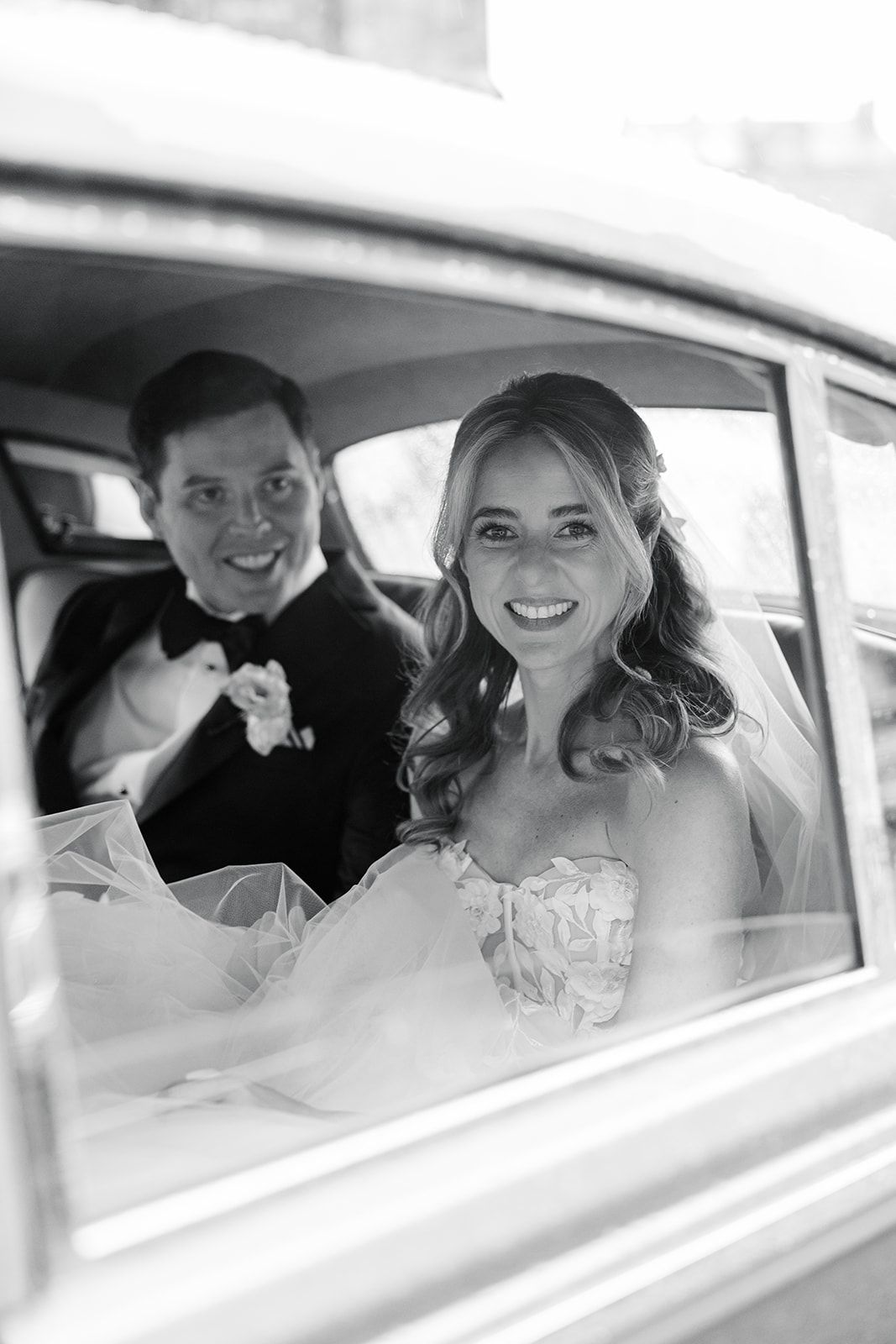 Black and white photo showing joyful smiles through a car window during a wedding celebration.