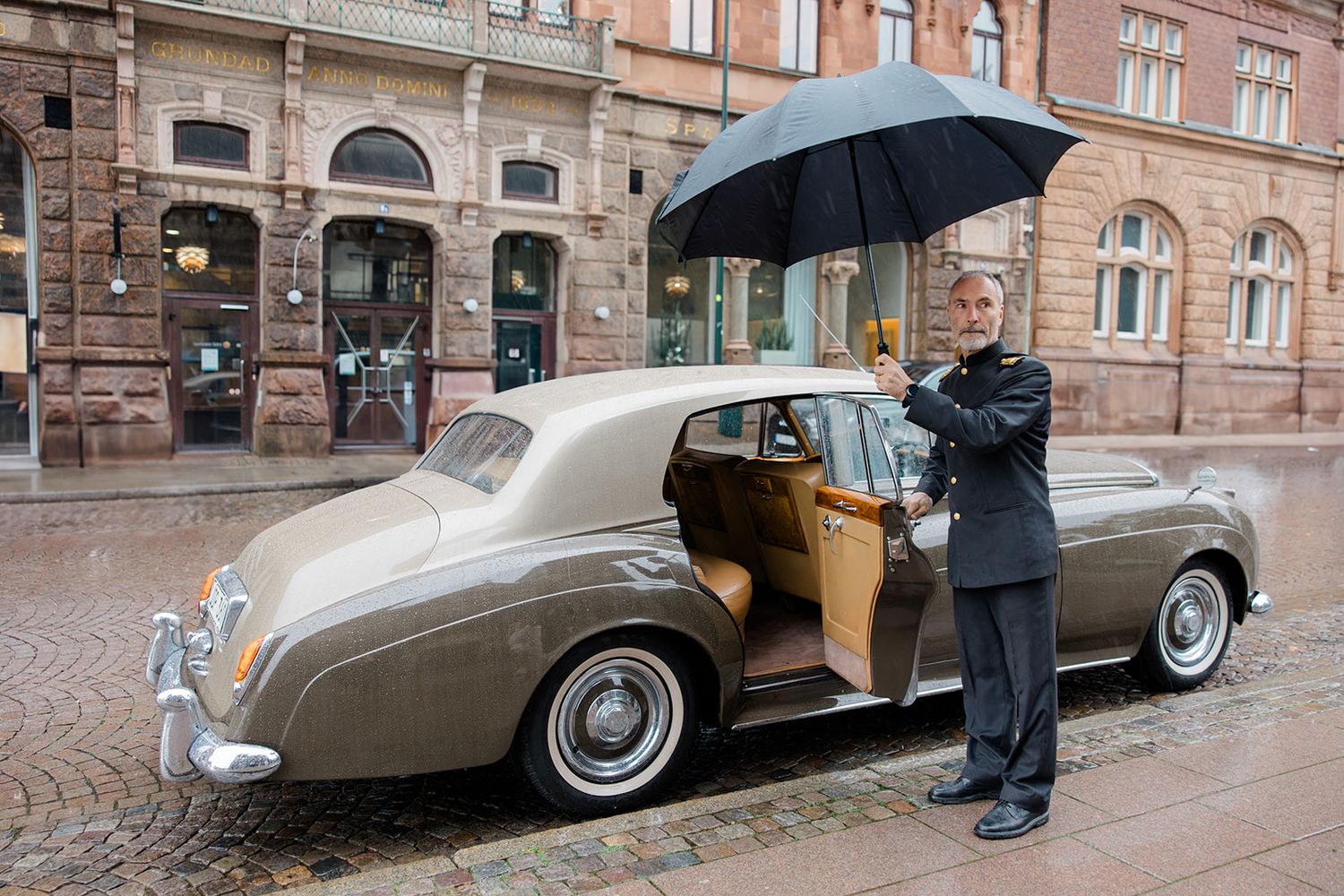 A vintage silver car parked on a cobblestone street with classic architecture in the background.