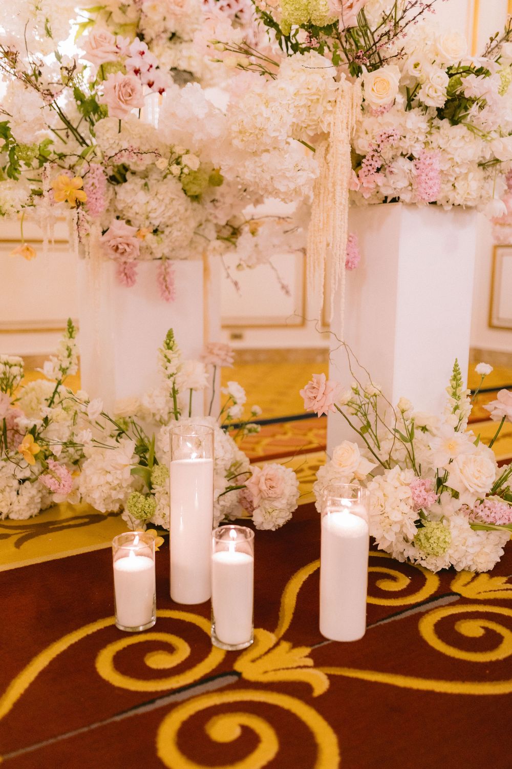 Elegant wedding reception decor with pink cherry blossoms and white candles arranged along a carpeted floor with gold swirls.