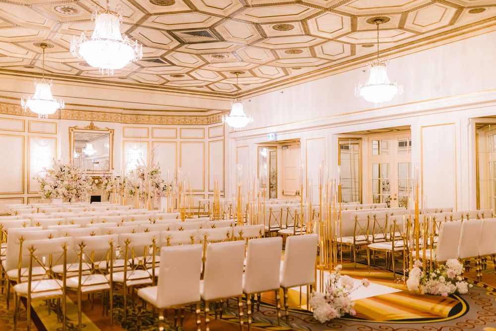 Elegant ballroom with ornate gold trim, crystal chandeliers and white chairs arranged in rows for a formal event.