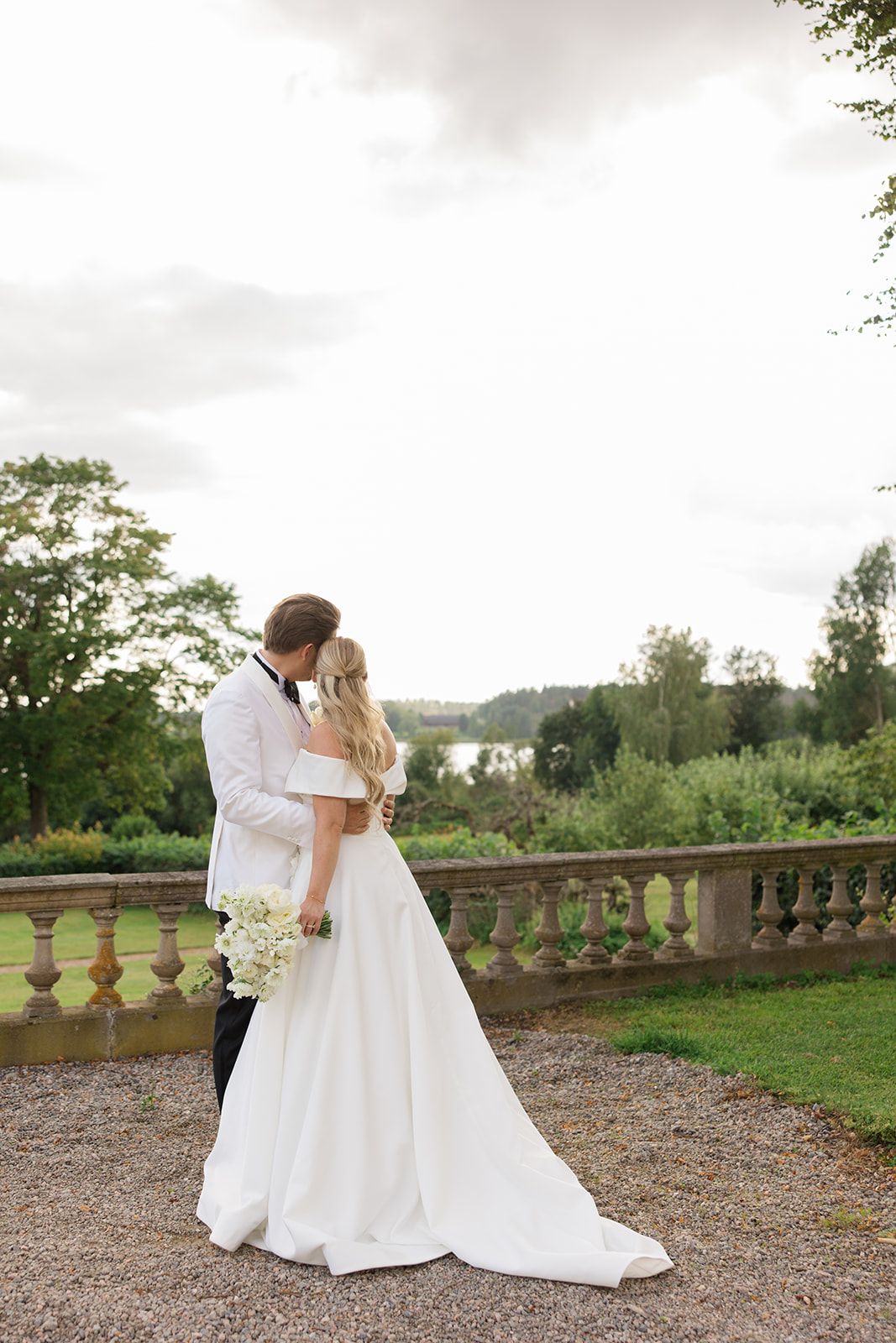 Two figures in white wedding attire embrace on a gravel terrace overlooking a scenic countryside landscape.