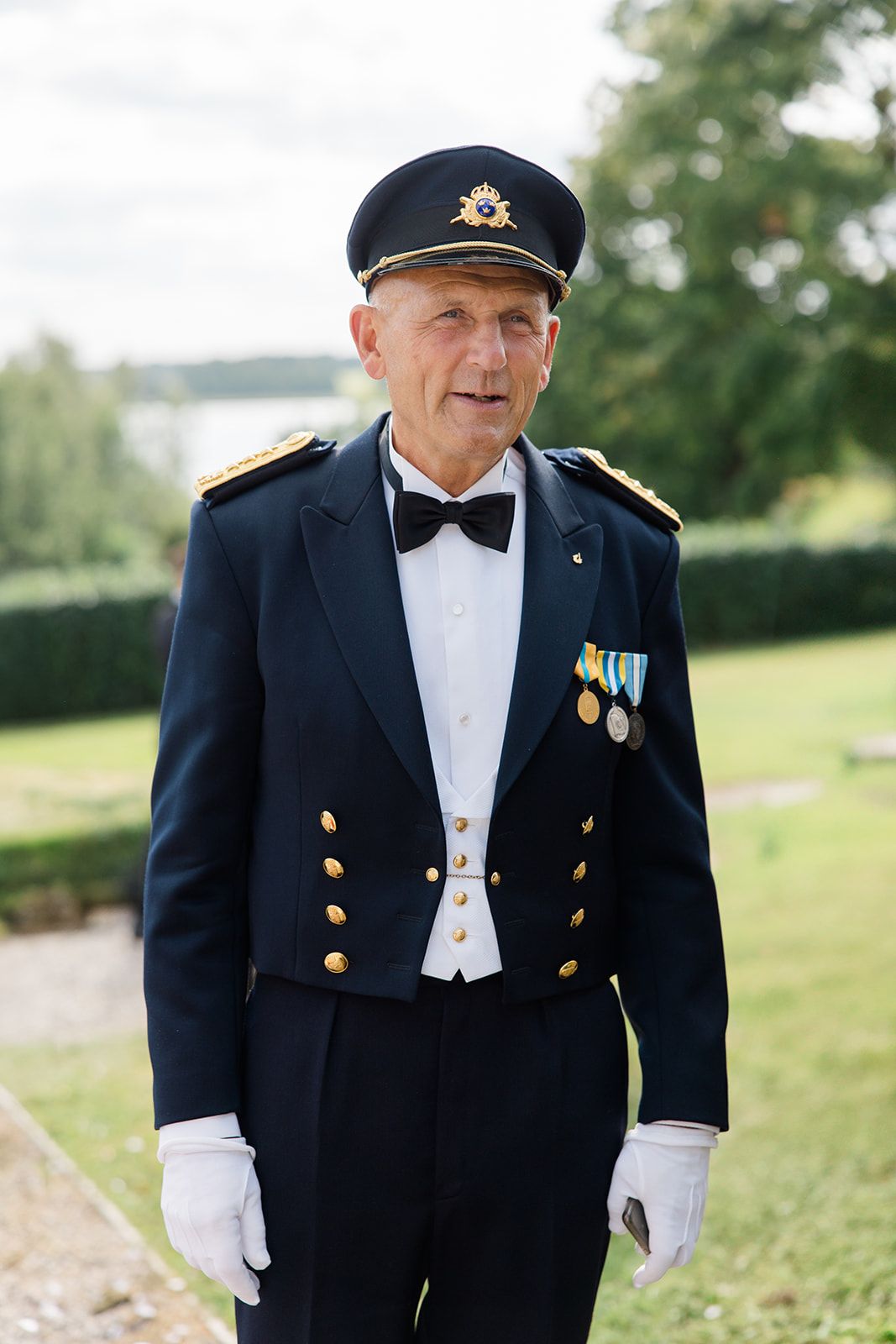 Military veteran in formal dress uniform with medals and white gloves stands outdoors in a garden setting.