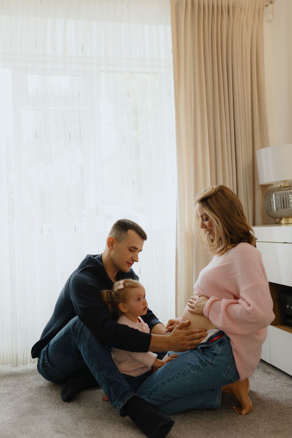 Family shares intimate moments together while relaxing on carpeted floor in bright, sunlit room.