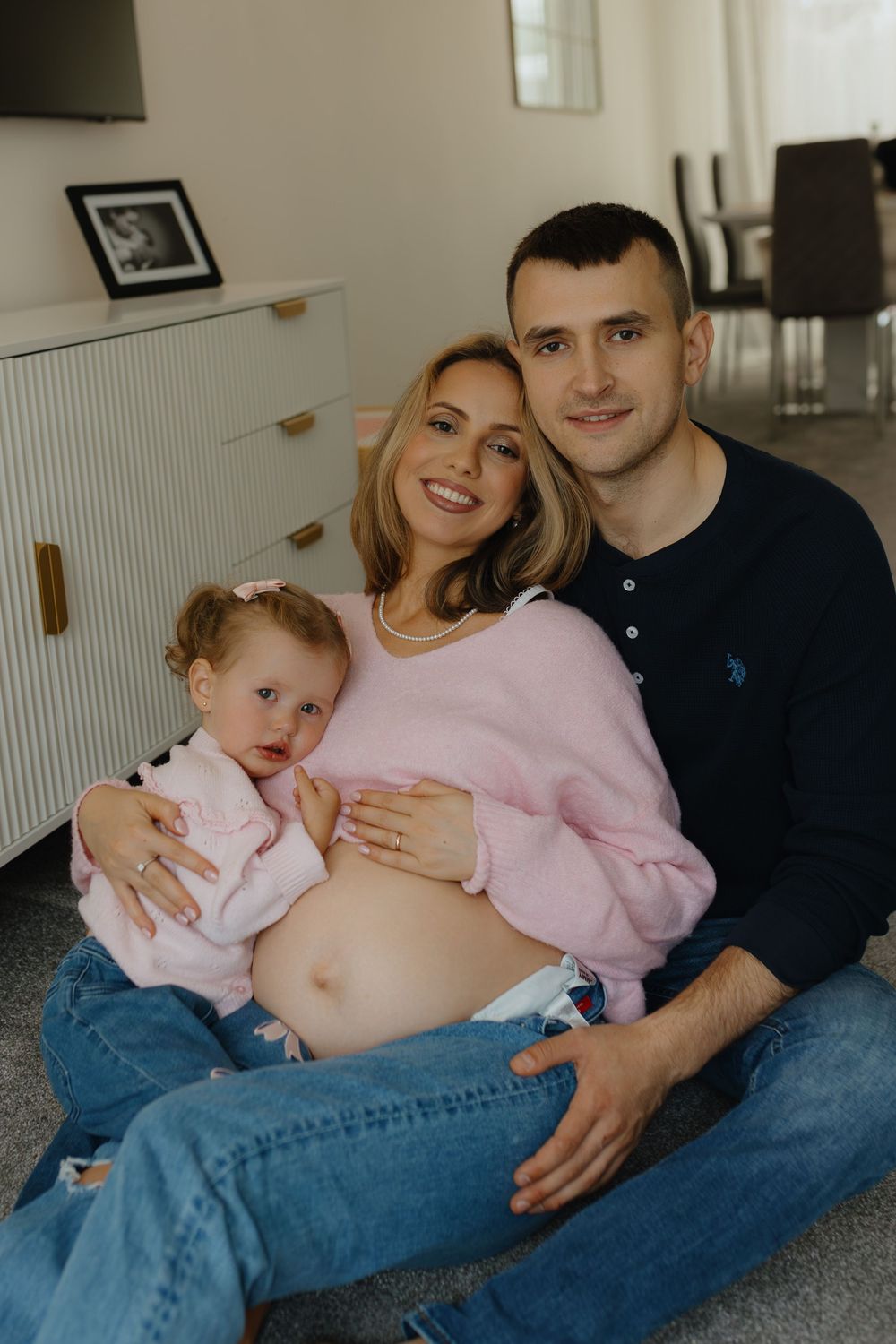 Family sits together on floor sharing tender moments with pregnant belly and young child in matching pink sweaters.