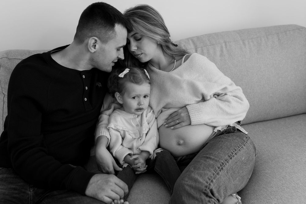 An intimate black and white family portrait on a couch showing a tender moment during pregnancy.