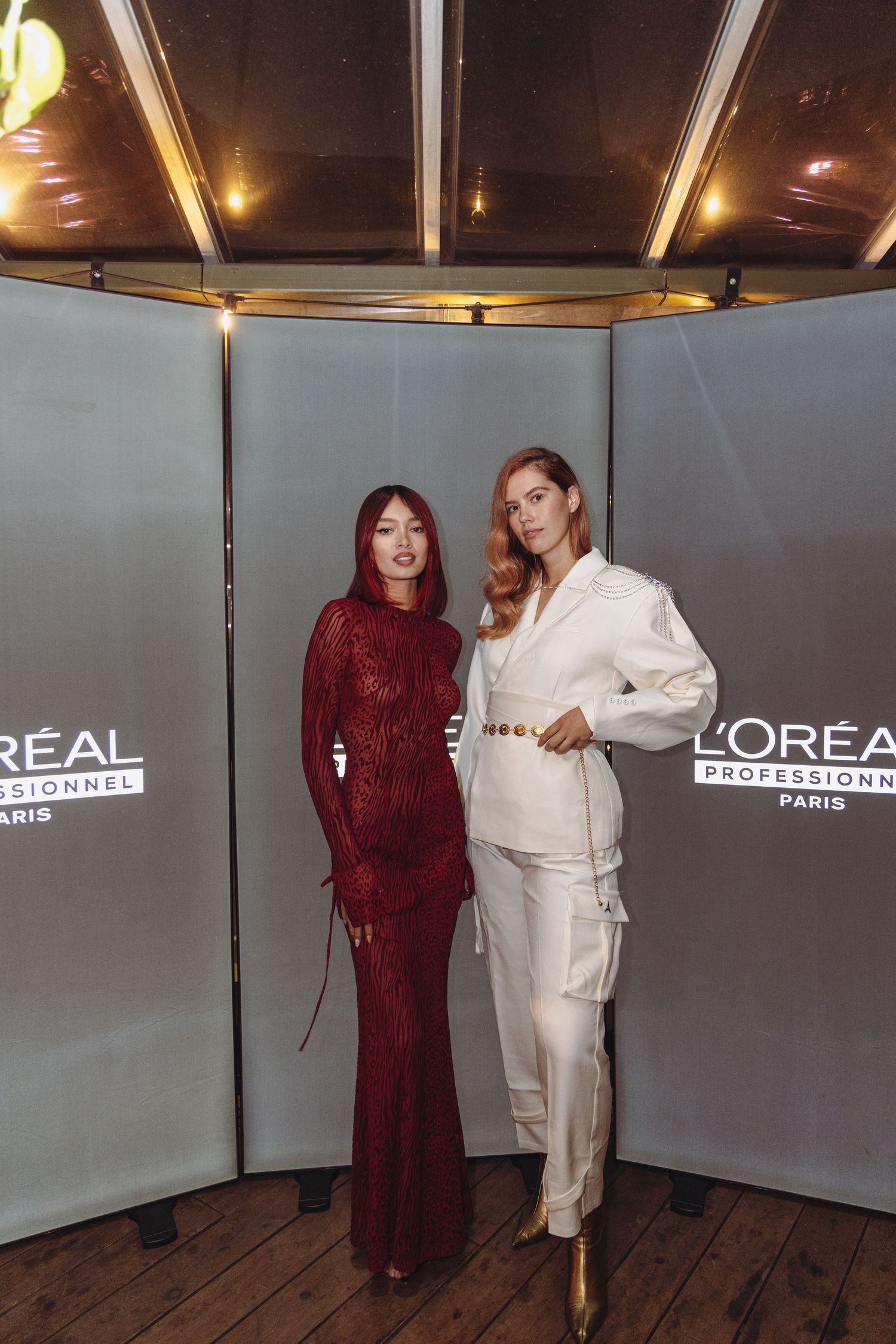 Two people pose at a L'Oreal Professional event, one wearing a burgundy sequined gown and another in white formal attire.