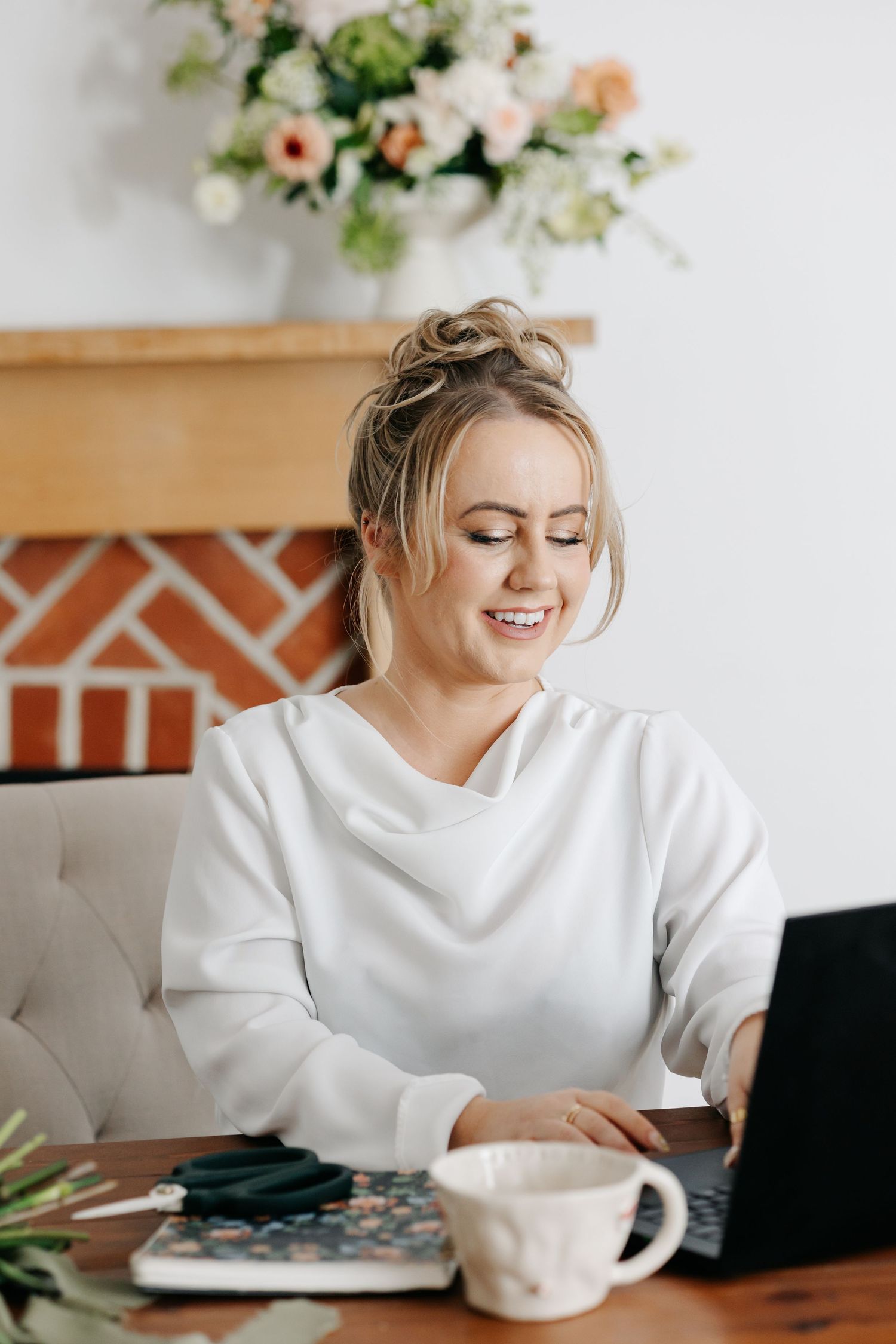 Woman in white sweater working from home at wooden desk with laptop, coffee cup and floral decor in background.