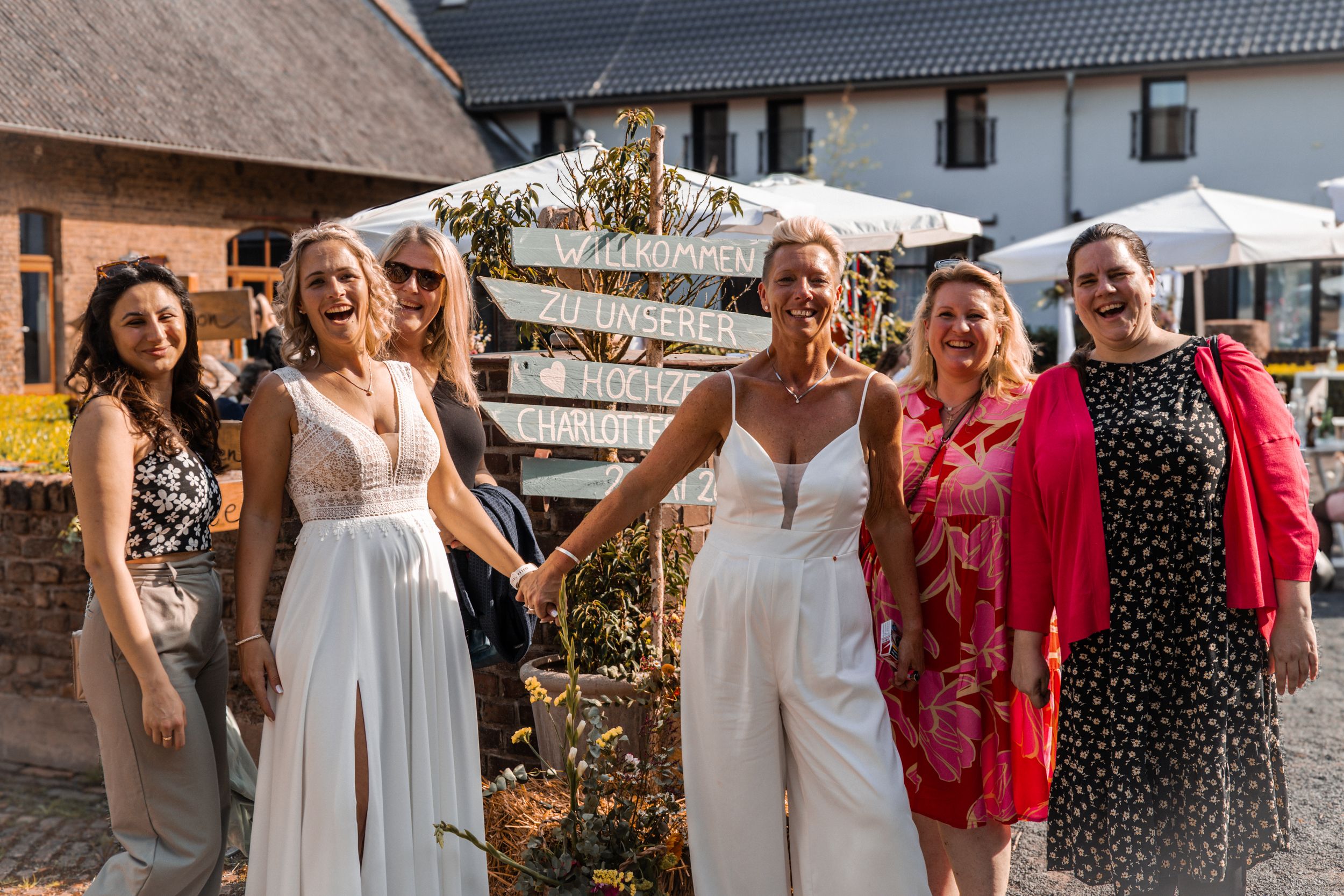 A group of people in summer attire gather outdoors for a festive celebration in front of a white building.