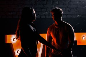 Two silhouetted figures illuminated by orange neon lighting against a dark brick wall background.