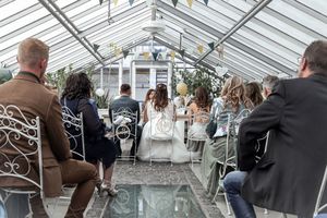 A wedding ceremony takes place inside a bright greenhouse with white chairs and floral decorations.