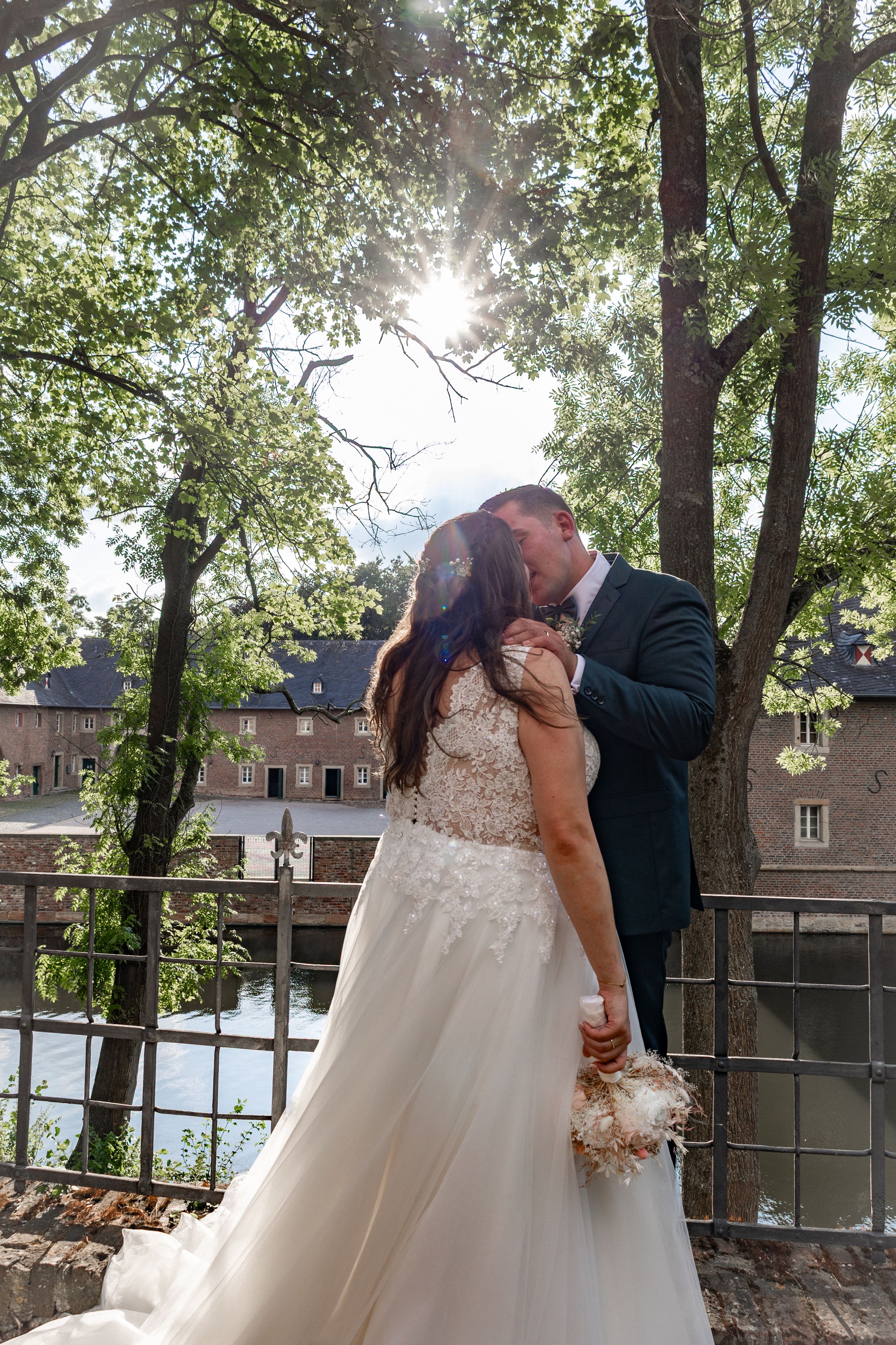 A couple in formal wedding attire shares a romantic kiss under sunlit trees in an outdoor garden setting.