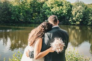 A couple in wedding attire embraces while standing near a serene lake surrounded by lush green trees.