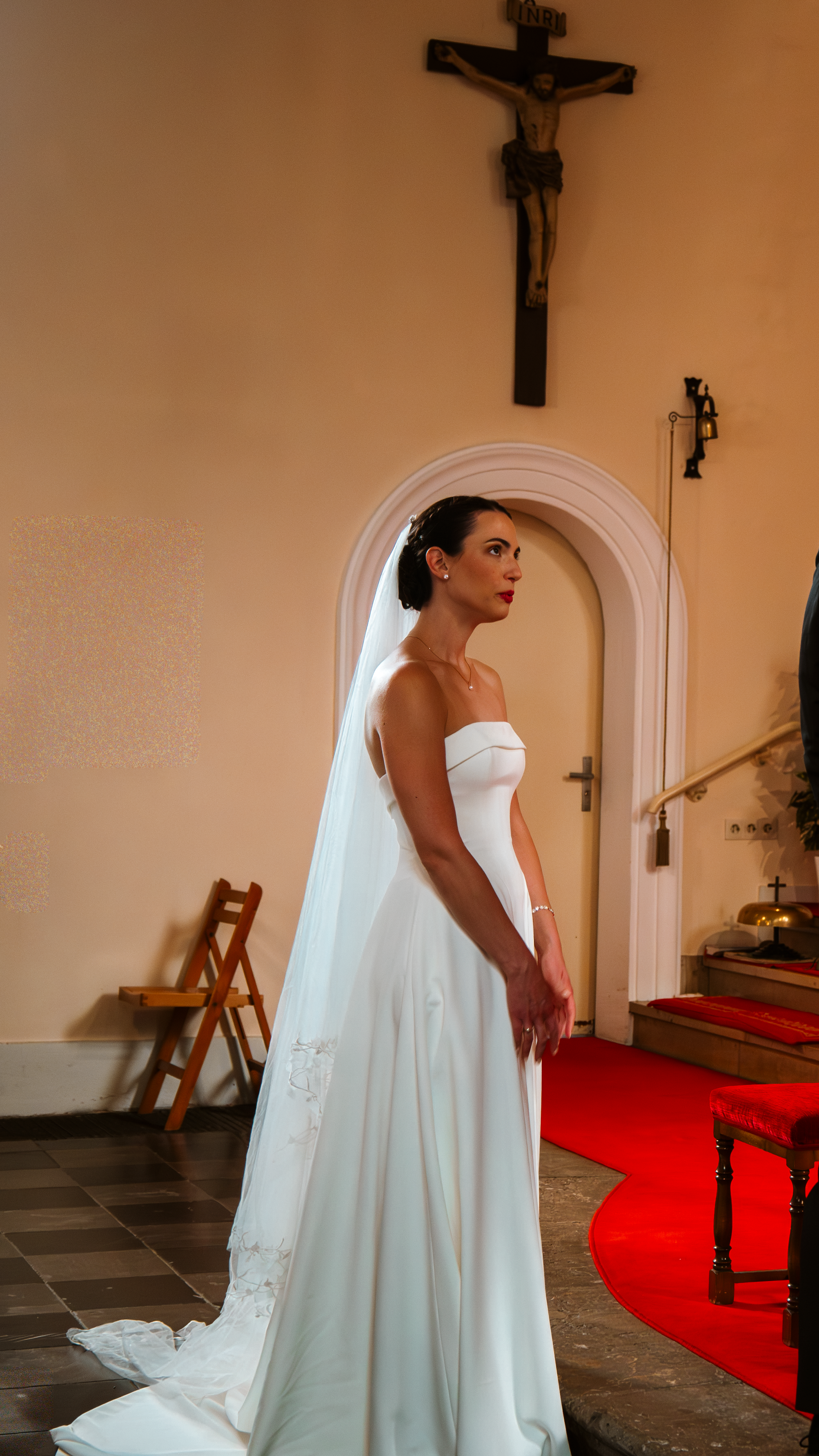 A bride in a flowing white wedding dress stands in a church interior with a red carpet and crucifix on the wall.