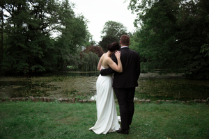 A couple in wedding attire embraces while standing on a lush green lawn surrounded by trees.