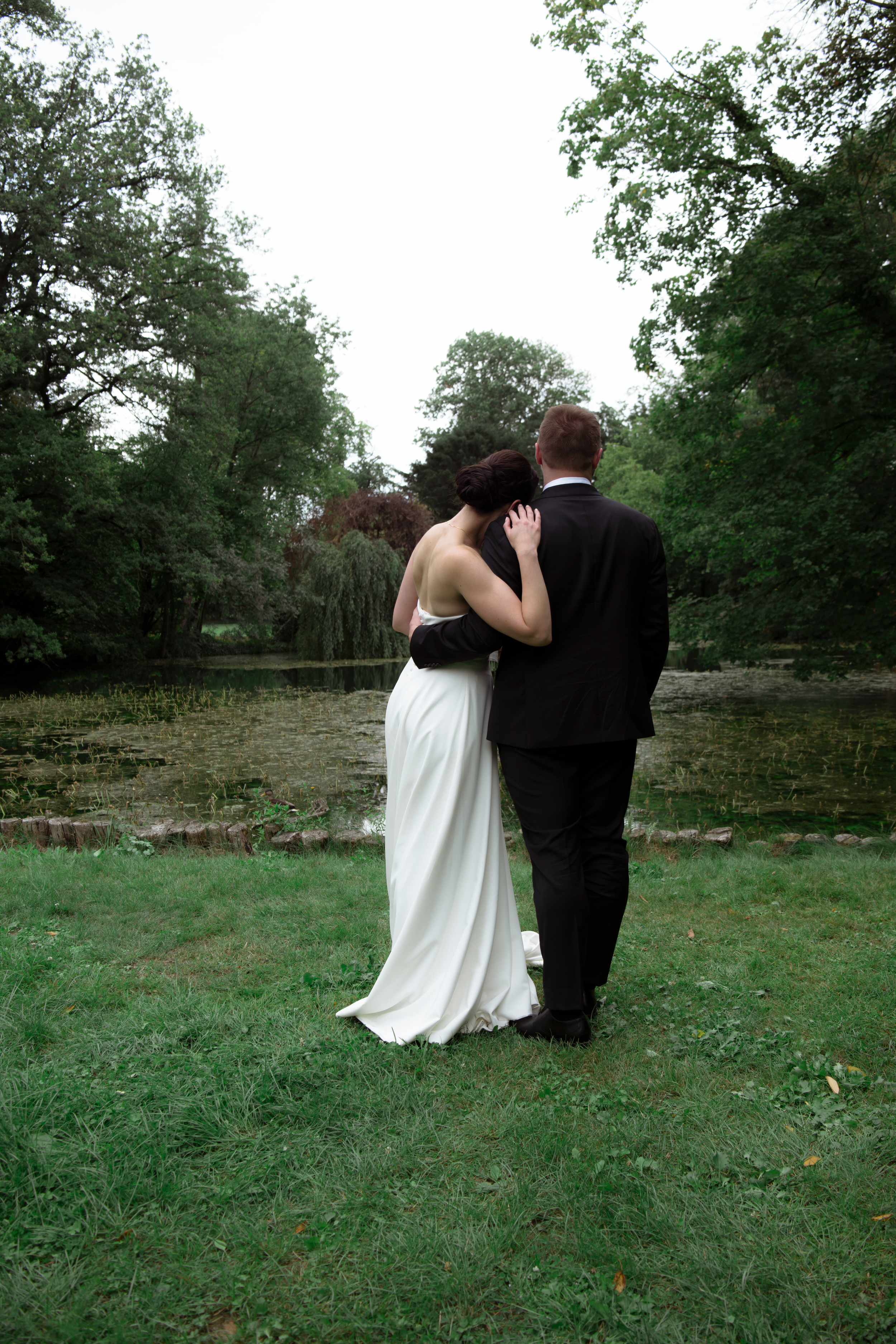 A couple in wedding attire walking away together across a green field surrounded by trees.