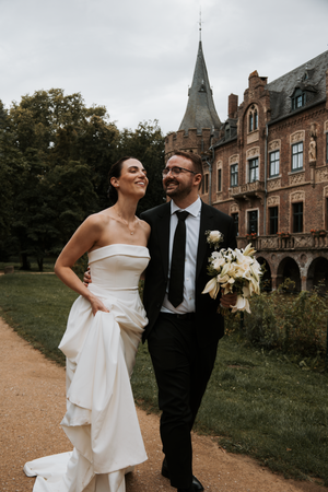 A romantic wedding couple laughs together while walking along a path near a historic brick mansion.