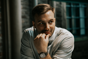 Professional portrait of someone in a white button-down shirt sitting in a modern office setting with dark lighting.