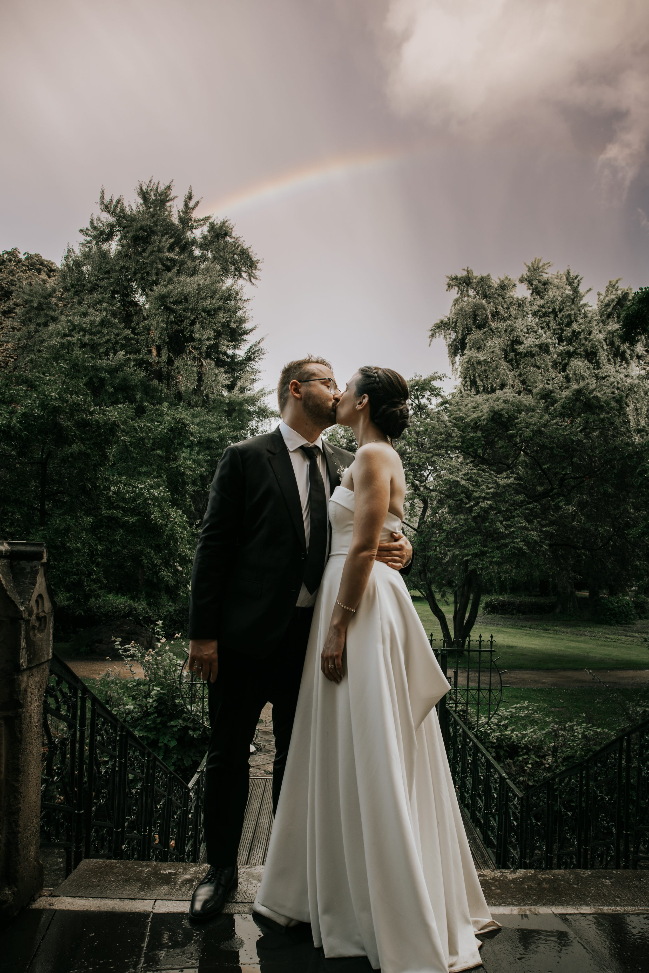 Romantic wedding couple shares intimate moments in an outdoor garden setting with dramatic cloudy sky backdrop.