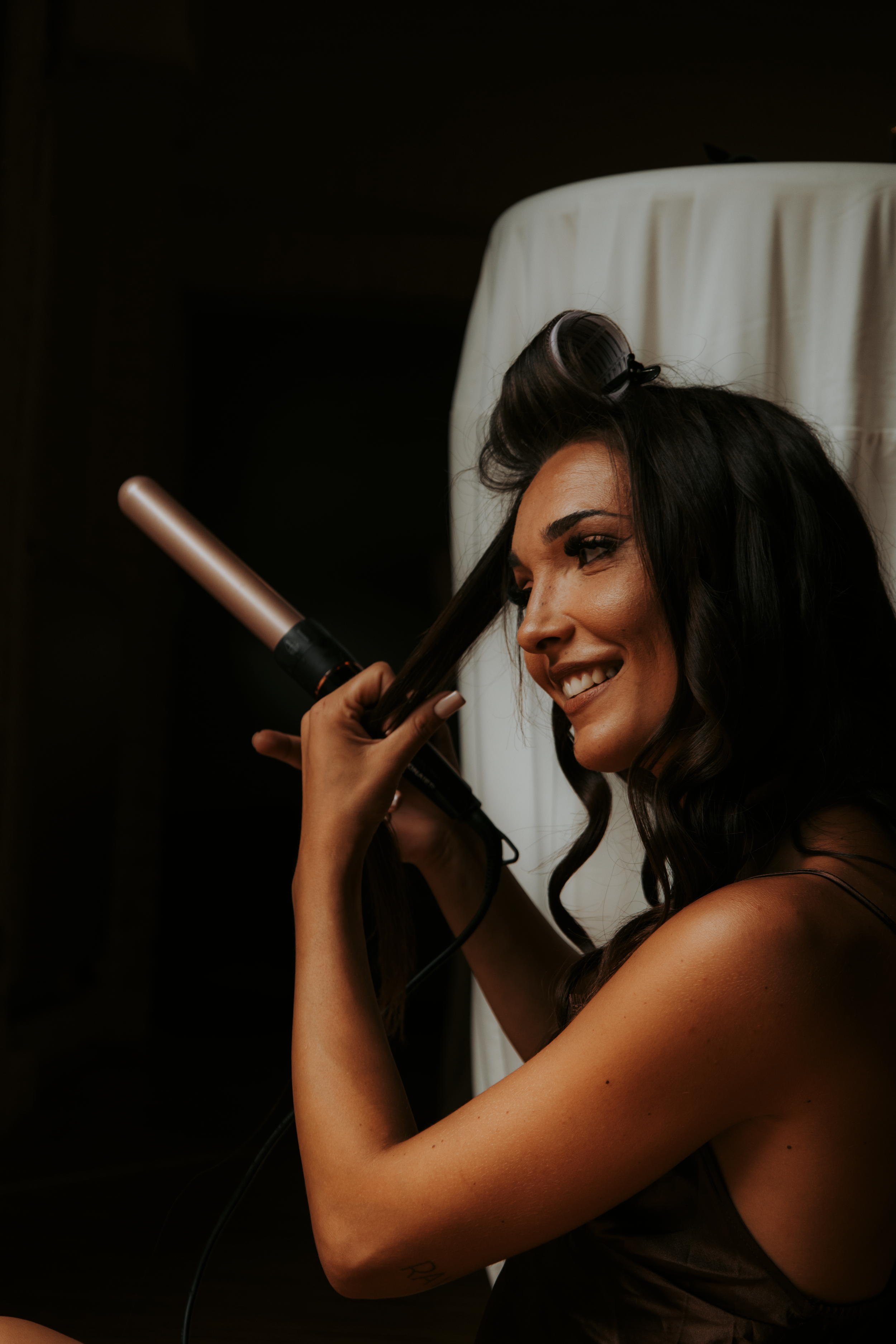 A smiling woman with dark curly hair uses a curling iron while getting ready, wearing a satin dress in warm lighting.