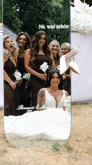 Bride in white lace gown sits before mirror with bridesmaids in brown dresses, tossing heels at outdoor wedding.