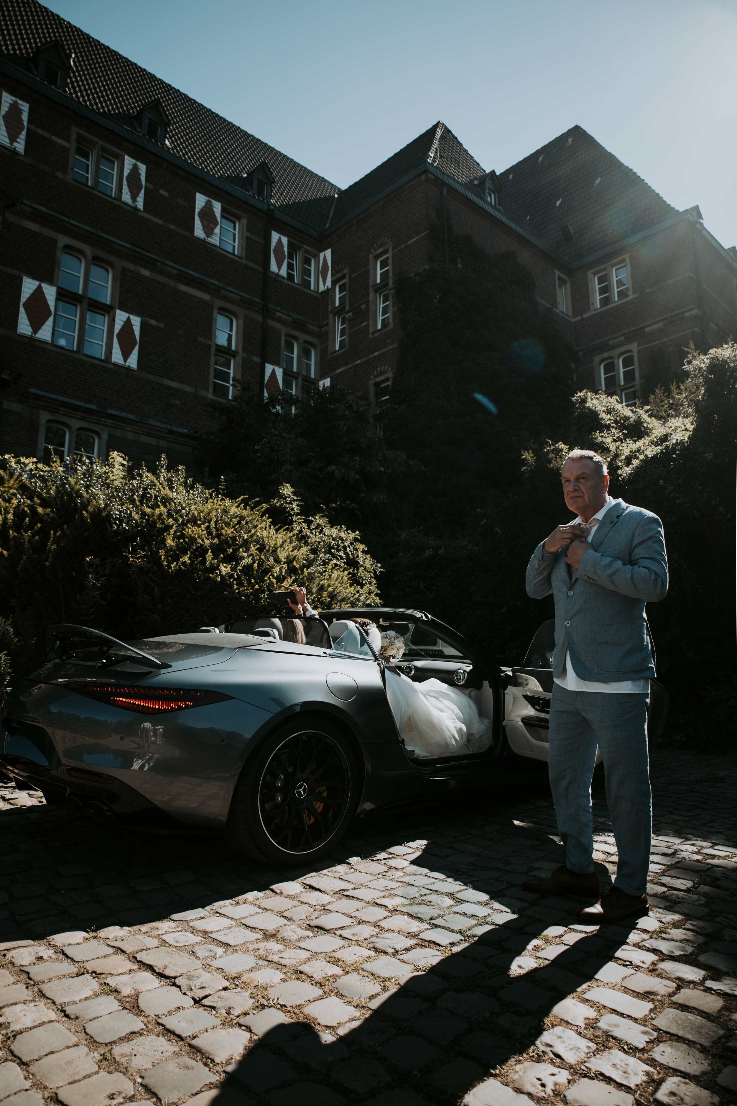 A man in a light blue suit stands beside a silver Porsche convertible on cobblestones outside a brick building.