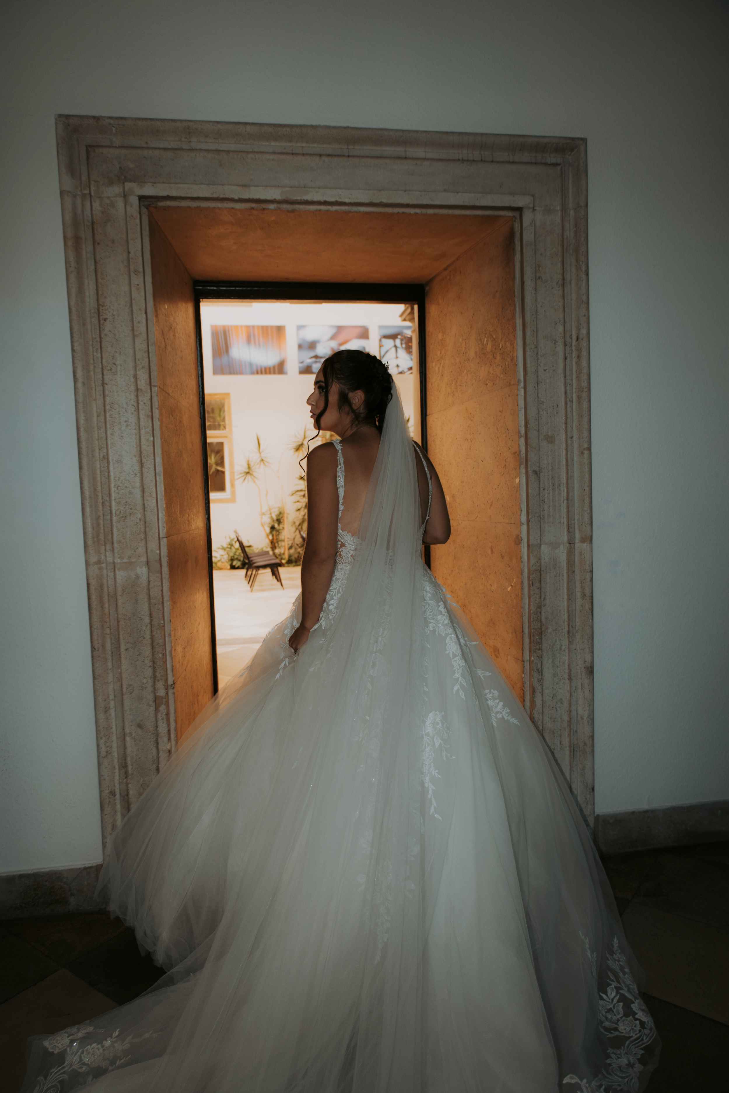 Bride in a flowing white tulle ball gown and veil stands in a stone doorway, backlit by warm golden light.
