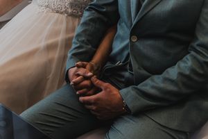 Person wearing dark teal jacket and pants sits with hands clasped in lap on white surface.