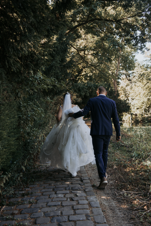 Bride and groom walking hand in hand down a cobblestone path surrounded by lush green trees on their wedding day.