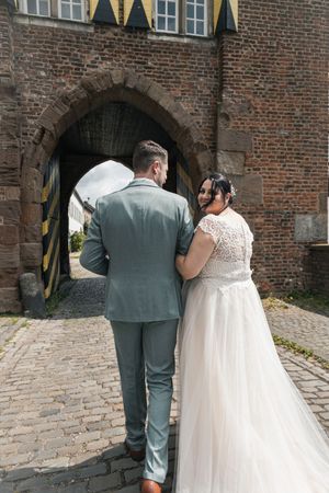 A couple in wedding attire walks along a cobblestone path beside a historic brick building with black and yellow banners.