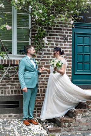 A couple in wedding attire dances playfully in front of a brick building with teal doors and white flower petals on the ground.