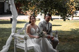 Couple sits together on white garden bench during outdoor wedding ceremony in sunlit garden setting.