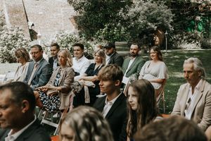 Wedding guests seated outdoors in rows of white chairs during a garden ceremony on a sunny day.