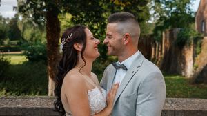 A couple in wedding attire share an intimate moment outdoors in a garden setting with trees in the background.