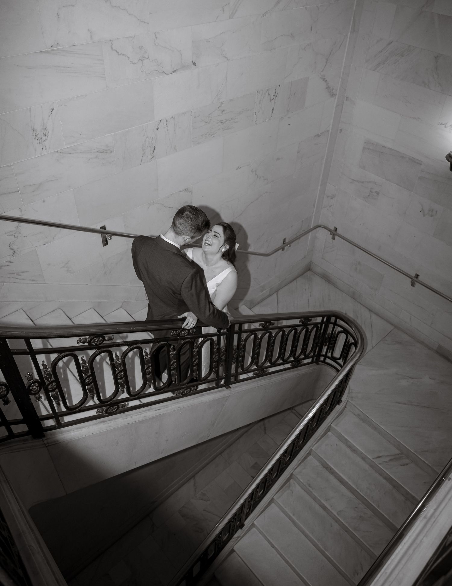 A couple shares a joyful moment by an elegant leaded glass window in a light stone interior hallway.