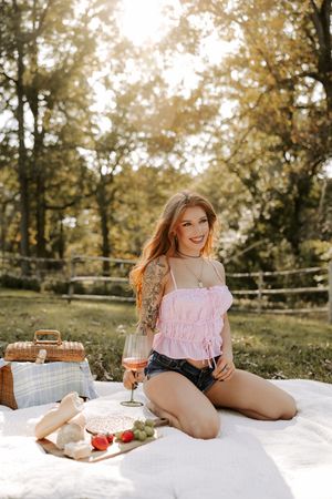 Outdoor picnic scene with pink top, white blanket, and rustic food spread in a sunny park setting.