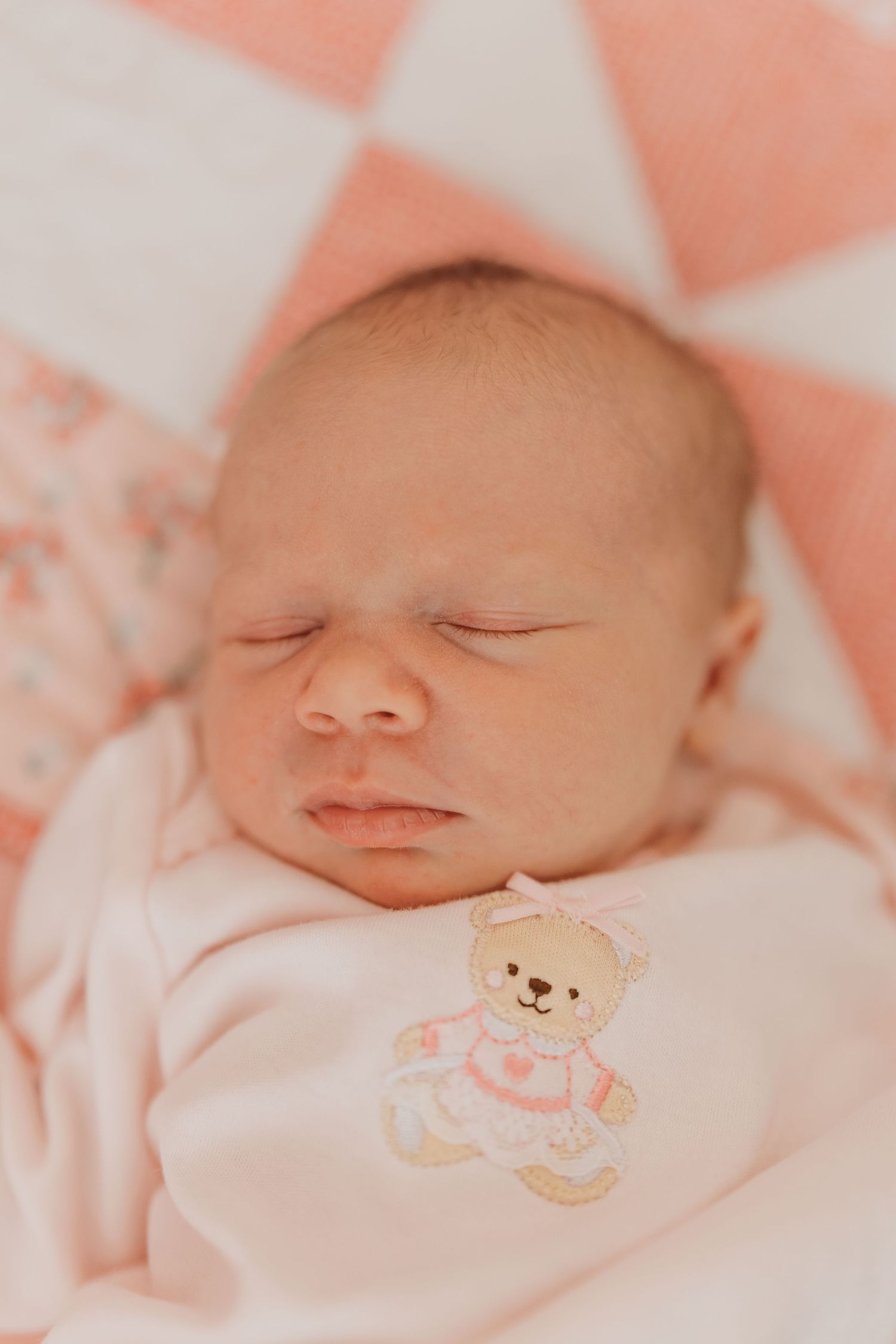 Sleeping baby wearing white outfit with teddy bear embroidery on pink bedding.