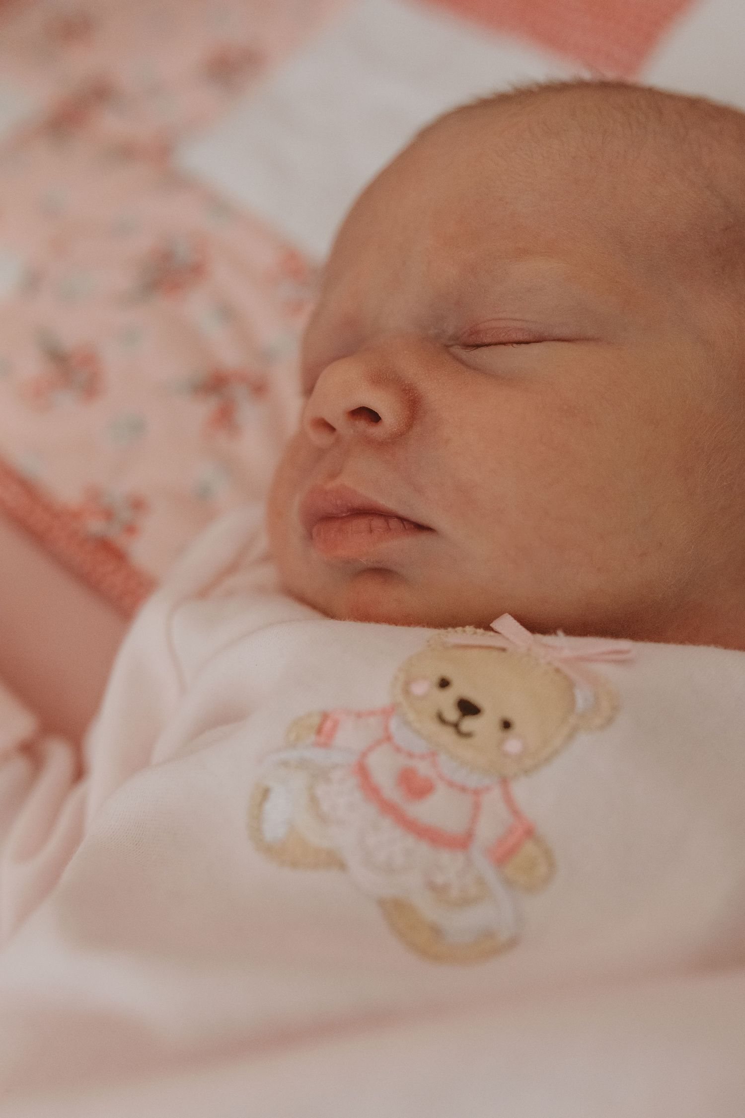 Close-up of sleeping baby on pink bedding with teddy bear detail on clothing.