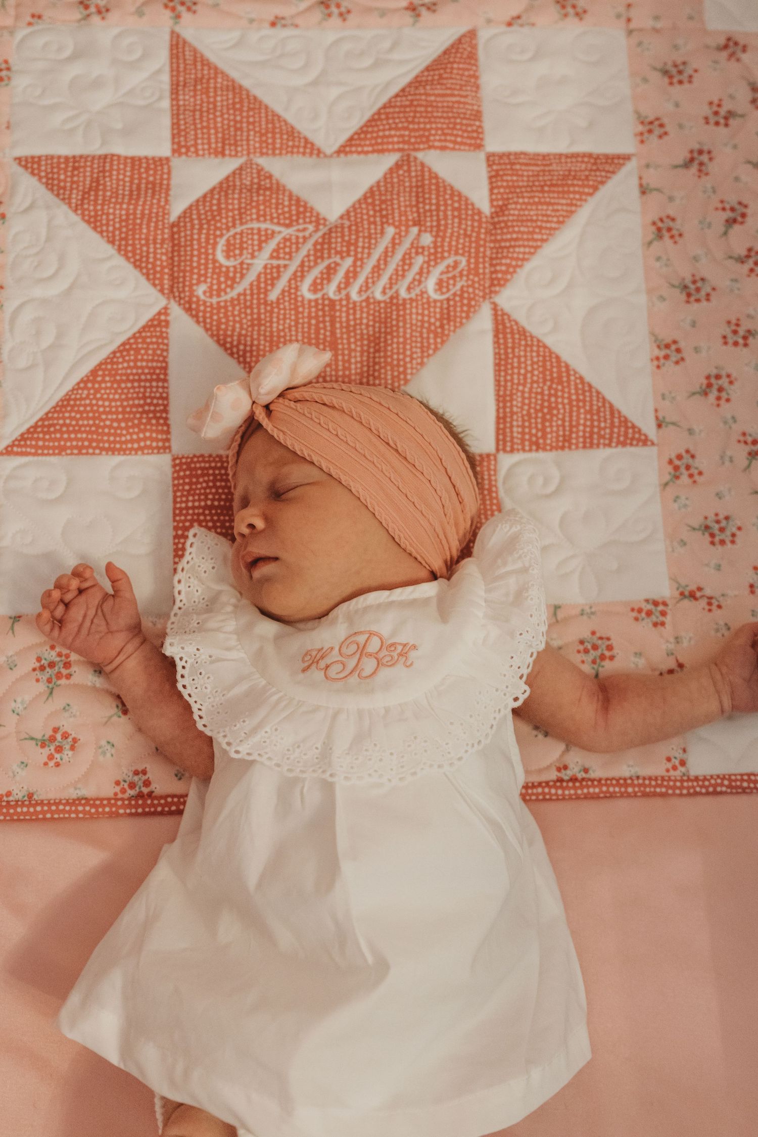 Baby in white dress resting on coral-colored quilt with embroidered name design.