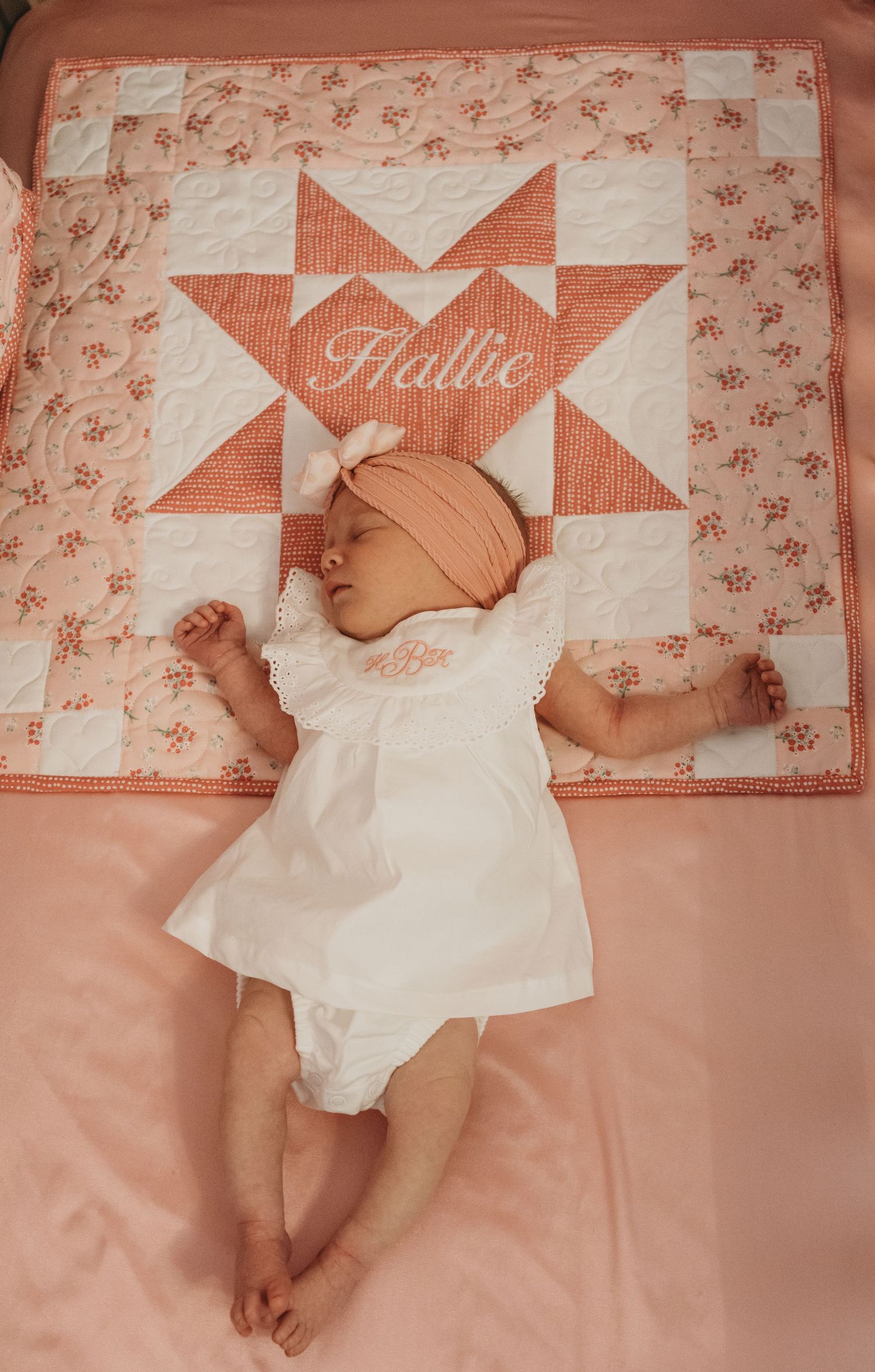 Newborn baby wearing white dress lying on pink quilt with geometric pattern.