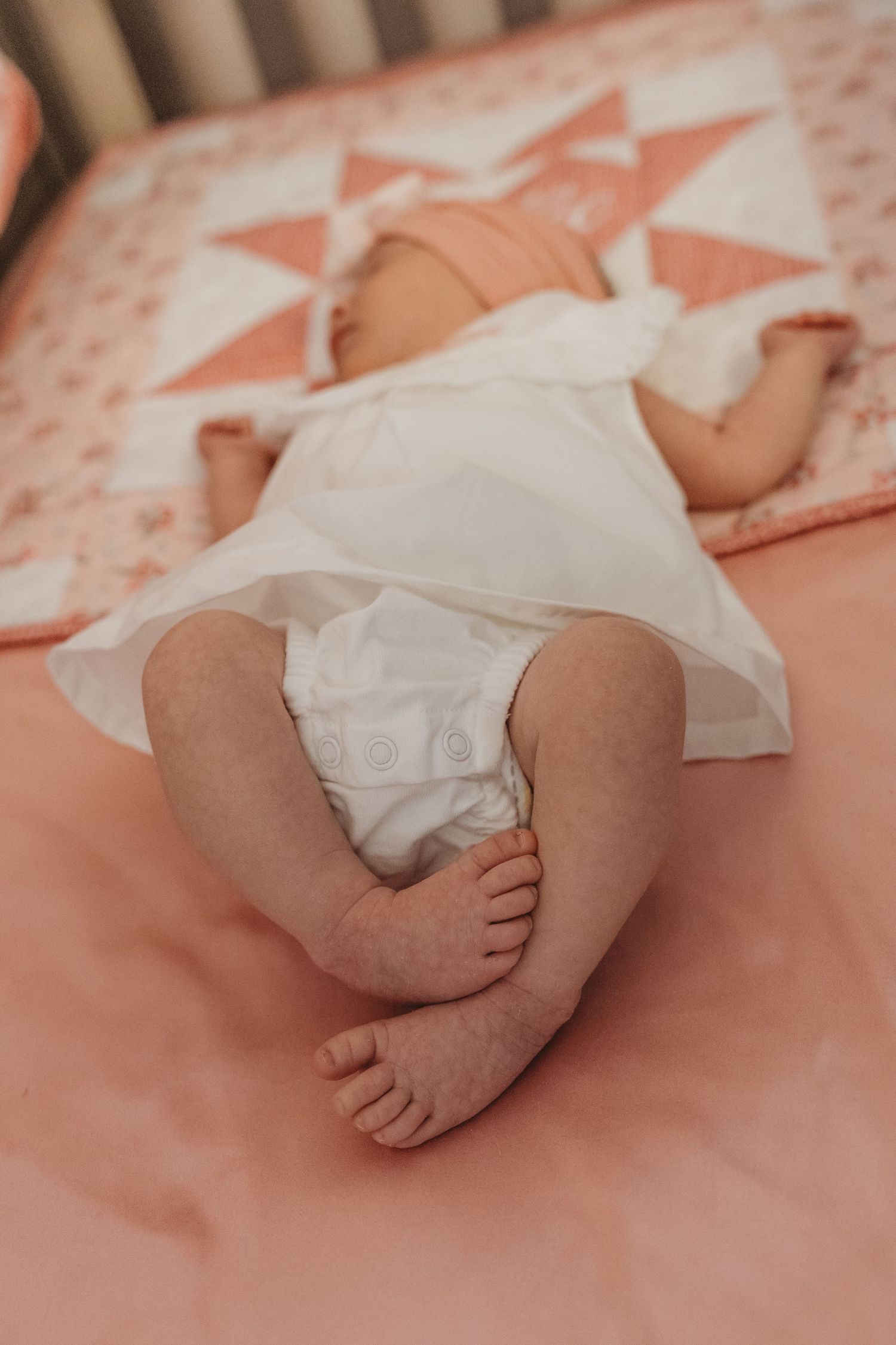 Close-up of baby feet and white dress against peachy pink bedding.