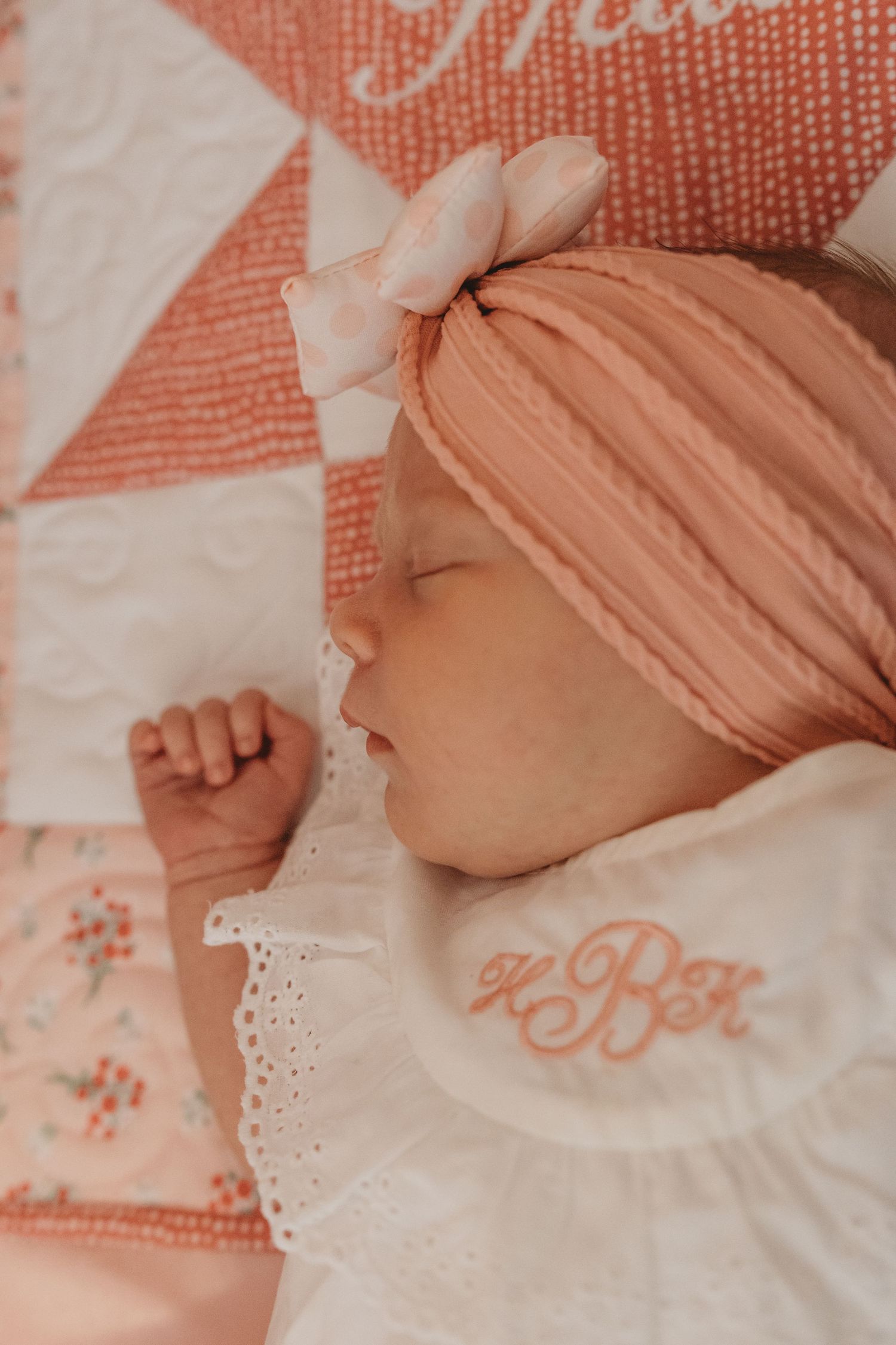 Detail shot of baby wearing white dress against coral-colored embroidered quilt.