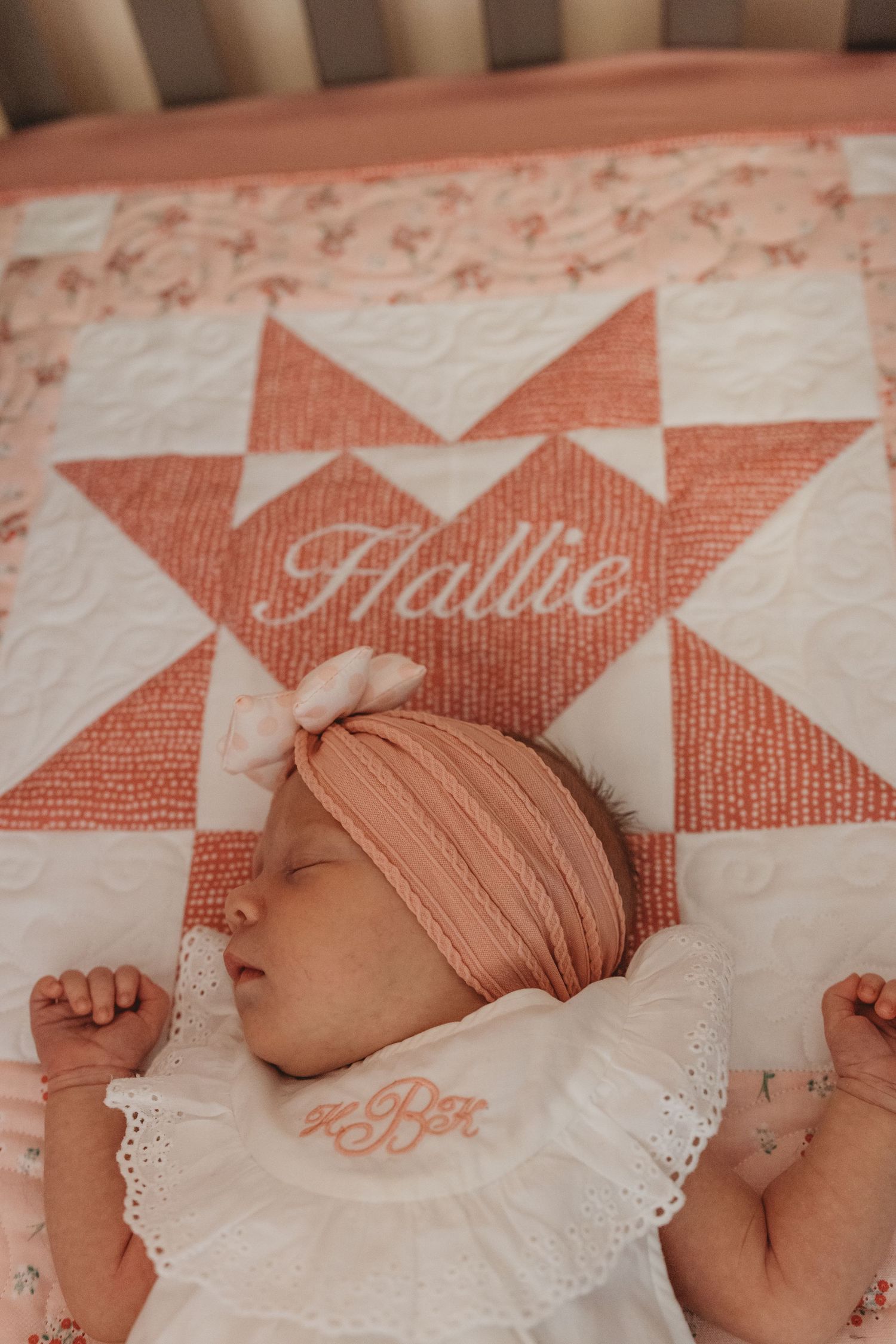 Sleeping newborn in white dress on decorative coral quilt with name embroidery.