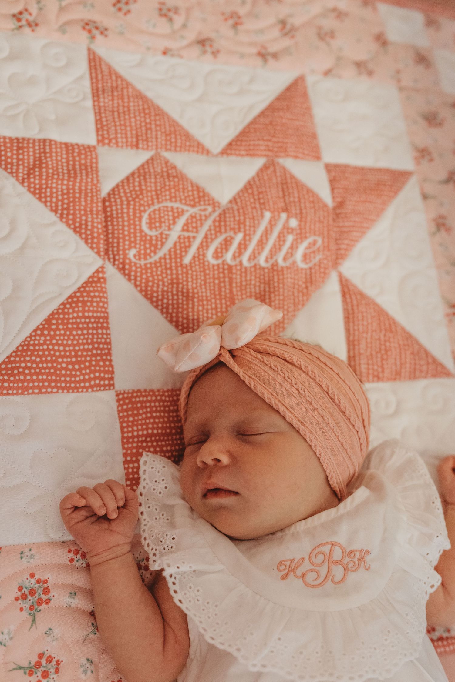 Newborn baby resting on a coral and white patterned quilt wearing a peach-colored cap.