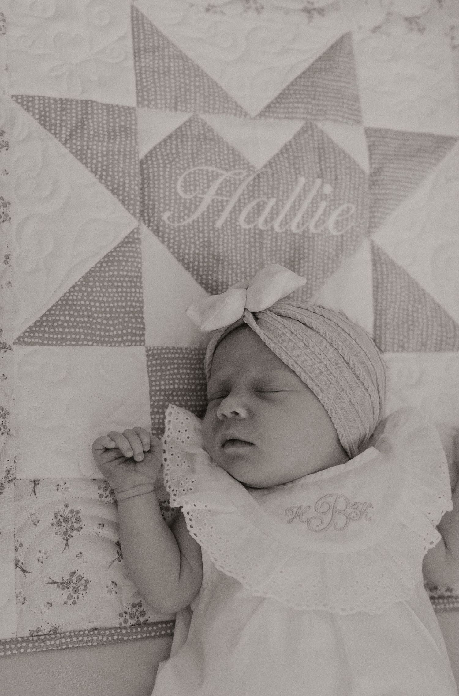 Newborn baby sleeping peacefully on a white and gray patterned quilt wearing a soft knit cap.