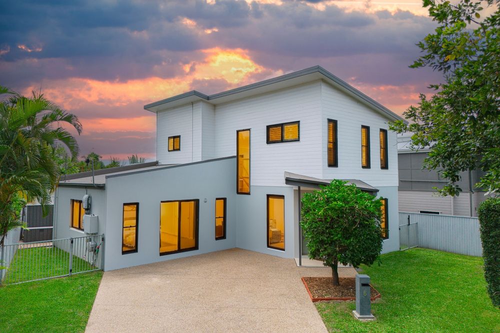 Modern white house with illuminated windows glows warmly against a dramatic sunset sky in a residential neighborhood.
