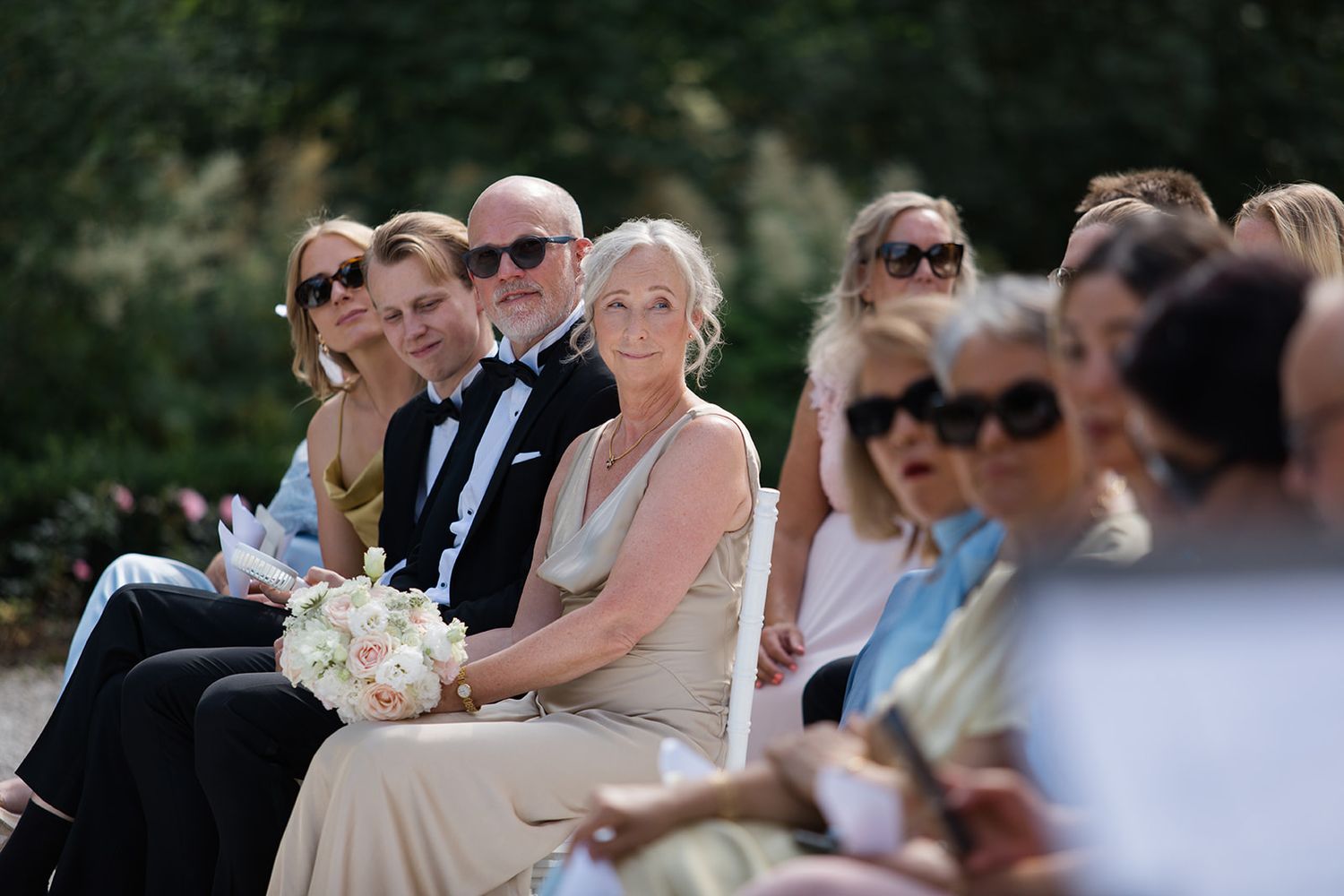 Wedding guests seated outdoors at a garden ceremony watch as the celebration unfolds on a sunny afternoon.